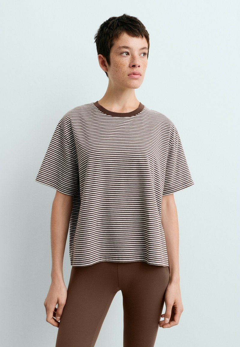 Jeune personne aux cheveux courts et foncés avec des taches de rousseur, portant un T-shirt rayé marron et blanc et des leggings marron unis sur un fond uni.