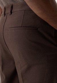 Pantalons marron ajustés en tissu texturé, avec une poche arrière épurée et des détails de couture subtils.