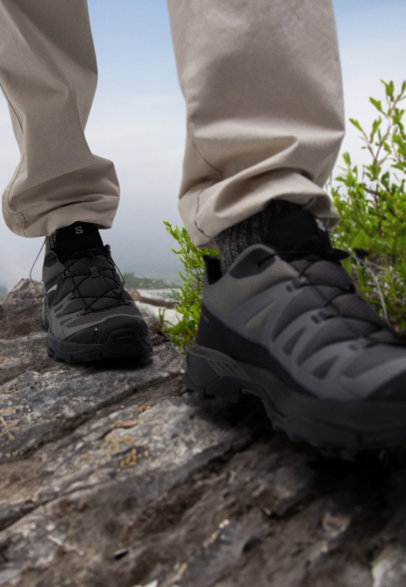 Czarne buty terenowe z teksturowaną cholewką, systemem sznurowania i solidną podeszwą, umieszczone na rocky powierzchni z zielenią w pobliżu.