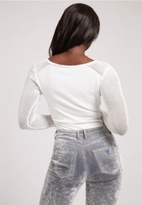 Maglia bianca a maniche lunghe con inserti in rete su spalle e braccia, abbinata a jeans grigi in velluto con tasche posteriori e dettaglio logo a triangolo.