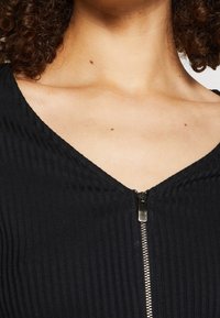 Top nero a coste con scollatura a V e una zip argentata sul davanti. Il tessuto ha una leggera texture ed è aderente attorno al collo.