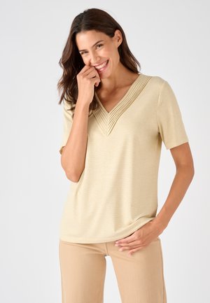 Lachende vrouw met donker haar draagt beige kortemouwer top met V-vormige geplooide halslijn en bijpassende beige broek tegen een effen achtergrond.