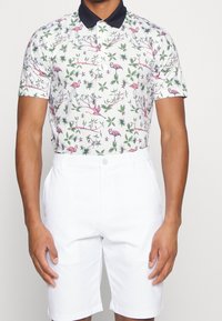 Polo à manches courtes avec une base blanche, arborant un motif coloré de flamants roses et de feuilles. Associé à un short blanc uni.