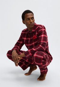Κόκκινη και μαύρη πιτζάμα καρό με κουμπιά, με τσέπη που φέρει εμφανές λογότυπο. Κατασκευασμένη από απαλό ύφασμα, σχεδιασμένη για άνεση.
