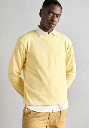 Felpa di cotone gialla con collo a giro, maniche lunghe e un logo ricamato discreto. Indossata sopra una camicia bianca con colletto.