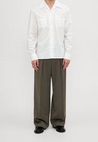 Vit skjorta med krage och två bröstfickor, kombinerad med löst sittande, khakifärgade byxor och svarta skor, på en neutral bakgrund.