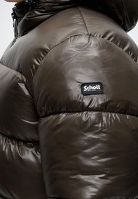 Gros plan sur la manche d'une doudoune marron foncé avec le patch logo Schott New York NYC visible sur le haut du bras.
