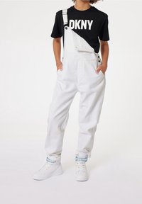 Barn som bär vita hängselbyxor med en rem undone och en svart DKNY-t-shirt, med händerna i fickorna, står i vita sneakers med blå DKNY-remmar.