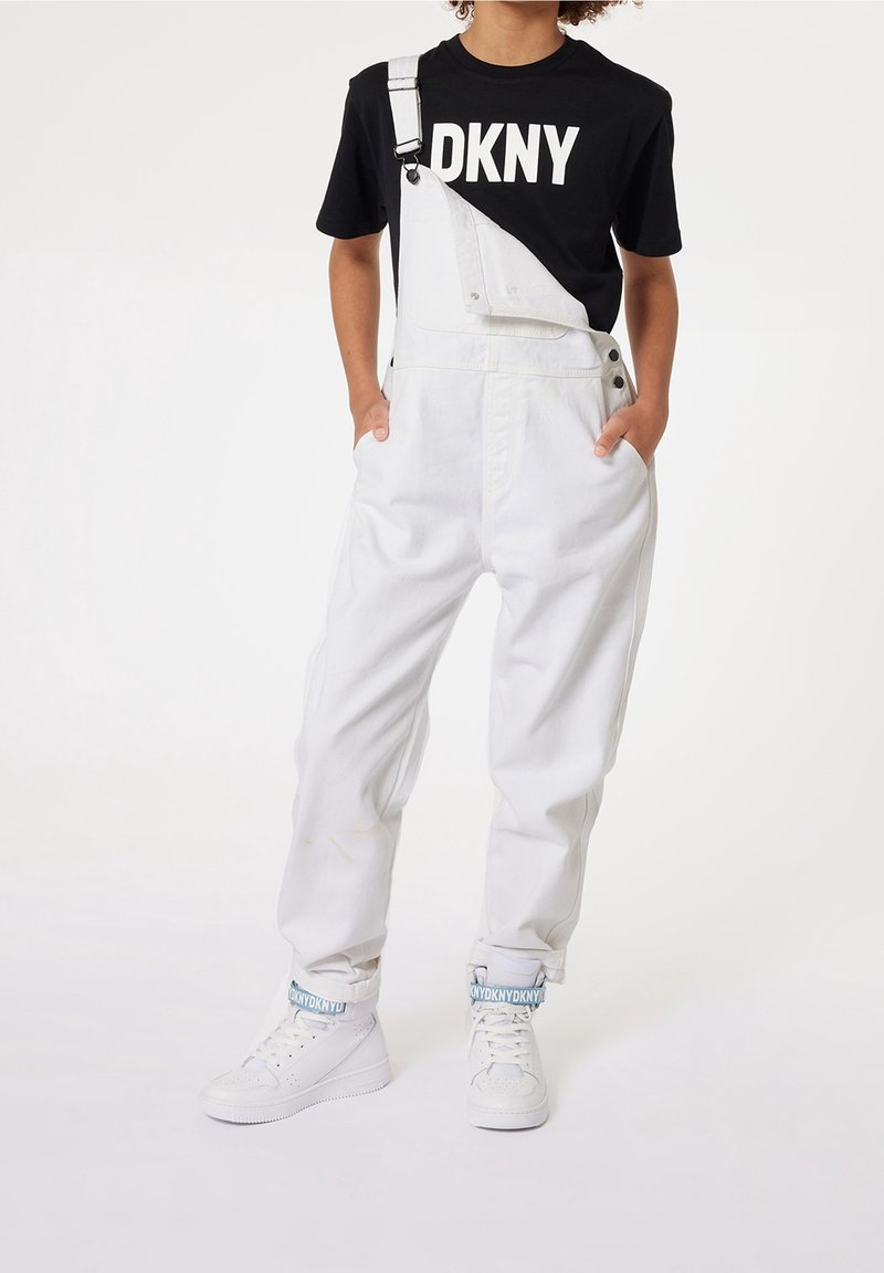 Barn som bär vita hängselbyxor med en rem undone och en svart DKNY-t-shirt, med händerna i fickorna, står i vita sneakers med blå DKNY-remmar.