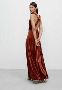 Long, sleeveless, rust-colored velvet dress with an open back, tied straps, and a flowing shape. Soft texture and elegant drape.