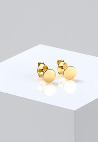 Pendientes de oro en forma de aro, con una textura suave y un acabado pulido. Montados sobre una superficie reflectante.