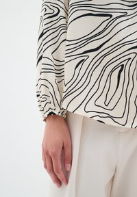 Lett blouse med et bølget sort linjemønster på en cremet baggrund, med samlede ærmer og en afslappet pasform, vist med cremede bukser.