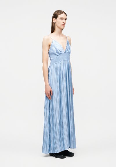 Γυναίκα που φοράει ένα μακρύ, ανοιχτό μπλε πλισέ φόρεμα με λεπτές τιράντες και βαθύ V-λαιμόκοψη, στέκεται μπροστά σε μια απλή λευκή φόντο.