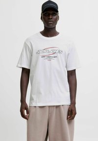 Melns cilvēks, ģērbies baltā Jack & Jones grafiskā T-kreklā, gaišos biksēs un melnā Pasadena cepurē, stāv uz vienkrāsainas baltas fona.