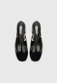 Black vegan patent leather shoes with a T-strap design, square toes, and a low block heel. Smooth texture with a glossy finish.