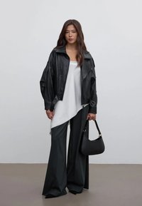 Zwarte leren jas met oversized pasvorm, wit asymmetrisch topje, grijze broek met wijde pijpen en een kleine zwarte handtas met gouden accenten.