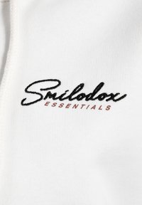 Witte stof met een gladde textuur, voorzien van het geborduurde logo "Smilodox Essentials" in zwart en een kleine rode accent.