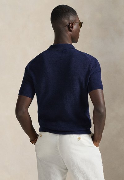 Πλεκτό πόλο μπλουζάκι σε ναυτικό μπλε χρώμα με κοντά μανίκια, με υφή επιφάνειας και κλασικό γιακά. Φορεμένο με ανοιχτόχρωμες βερμούδες.