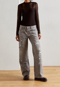 Schwarzes Langarmoberteil mit Punkten, kombiniert mit grauen weiten Jeans mit weißen Farbklecksen und übergroßen Fronttaschen. Braune Schuhe.