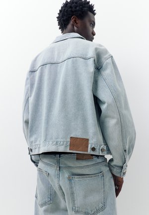 Osoba s krátkymi kučeravými vlasmi, oblečená v svetlomodrej rifľovej bunde a zodpovedajúcich džínsach, stojaca pred jednoliatym bielym pozadím.