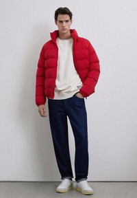Joven de pie con una chaqueta acolchada roja, sudadera crema, vaqueros azul oscuro y zapatillas blancas frente a un fondo liso.