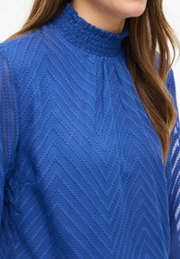 Blusa blu con motivo a zigzag, dotata di un alto collo smock, in tessuto testurizzato e maniche lunghe trasparenti.