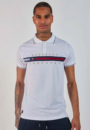 Homme tatoué portant un polo blanc avec le texte "Redskins Industry" et des rayures horizontales rouges et marine, se tenant devant un fond uni.