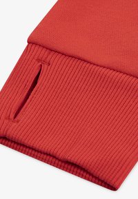 Material roșu cu nervuri, dotat cu un mansetă cu deschidere laterală. Textură moale, margini cusute cu grijă și un design strâns, ajustat.