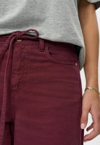 Kadar blizu osobe koja nosi visoko strukirane burgundske hlače s vezicom u struku i sivi majicu ubačenu unutar hlača, zlatnu narukvicu na zapešću.