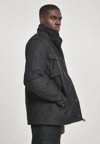 Black waterproof jacket with a high collar, multiple front pockets, and a zip closure. Features a relaxed fit and durable fabric texture.