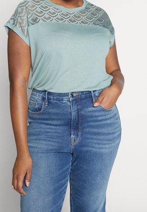 Vrouw draagt lichtgroen topje met kantpatroon en blauwe high-waisted jeans, met één hand in de zak en de andere ontspannen langs haar zijde.