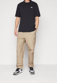 Černé polo tričko s krátkými rukávy a bílým logem Nike, doplněné béžovými cargo kalhotami a černo-bílými teniskami Nike. Zobrazený je ležérní styl.