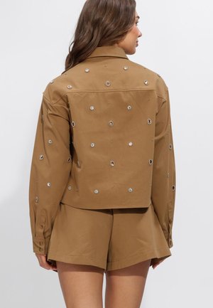 Frau trägt ein braunes Langarmshirt und passende Shorts, das Shirt ist mit kleinen runden silbernen Verzierungen dekoriert, die Frau ist von hinten zu sehen.