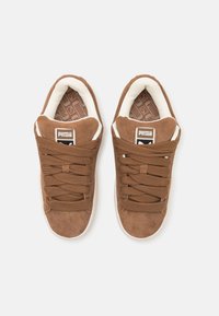 Brown suede sneakers featuring thick laces, a white padded collar, and a patterned insole. Logo displayed on the tongue.