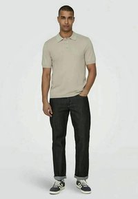 Camisa polo beige de manga curta com colarinho, combinada com jeans azul escuro e sapatilhas azul-marinho, encostada a um fundo claro.