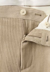 Pantalones de pana beige con costillas verticales, cierre con cremallera y dos botones en la cintura. Textura suave con bolsillos funcionales.