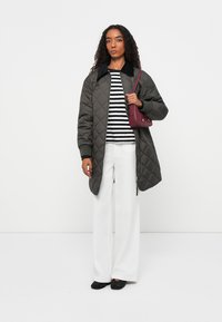 Gequiltte olijfgroene jas met een zwarte kraag, gestreepte zwart-witte blouse, witte wijde broek, en een bordeauxrode handtas, zwarte schoenen.
