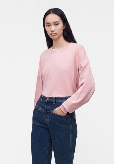 Femme aux longs cheveux foncés portant un sweat-shirt rose et un jean bleu, debout avec une main dans la poche devant un fond uni.