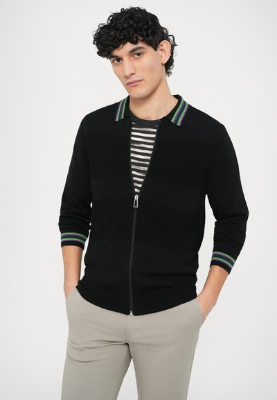 Čierny kardigán na zips s golierom s pruhmi v zelenej, fialovej a modrej farbe. Nosený cez sivé a biele pruhované tričko.