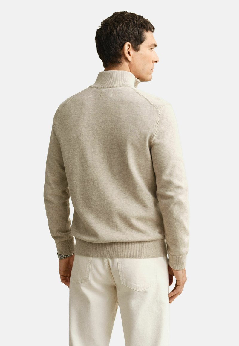 Hombre con cabello corto y oscuro, vestido con un suéter beige de cuello alto falso y pantalones blanco roto, visto desde atrás sobre un fondo liso.