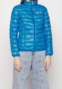 Niebieska puchowa kurtka z wysokim kołnierzem, zamkiem błyskawicznym oraz delikatnym, falistym pikowaniem. Posiada logo na piersi i gładki, błyszczący materiał.