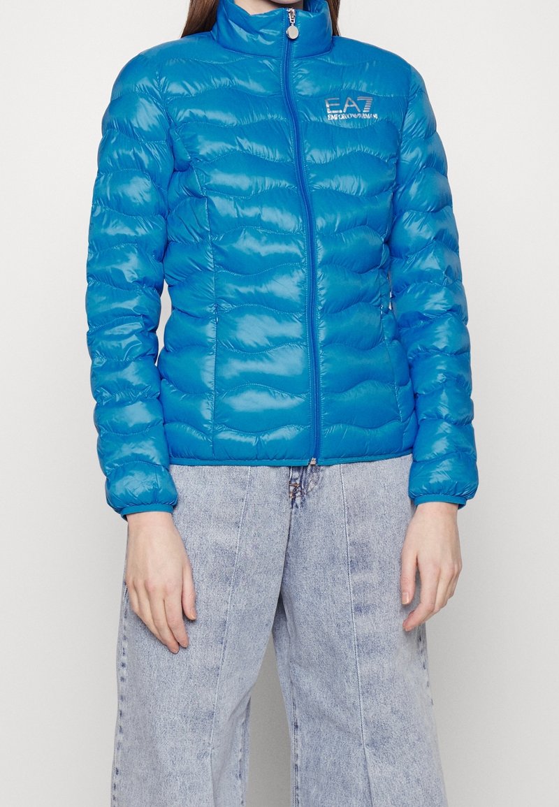 Niebieska puchowa kurtka z wysokim kołnierzem, zamkiem błyskawicznym oraz delikatnym, falistym pikowaniem. Posiada logo na piersi i gładki, błyszczący materiał.