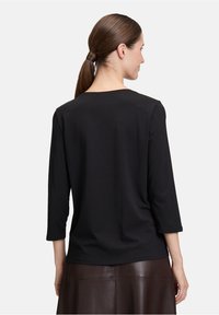 Vrouw met bruin haar in een staart, gekleed in een zwart top met driekwart mouwen en een bruine leren rok, gezien van achteren.