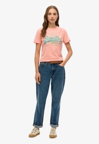 T-shirt en coton rose avec un texte vert indiquant "SuperDry", associée à un jeans bleu et des baskets beiges. Le modèle se tient devant un fond neutre.