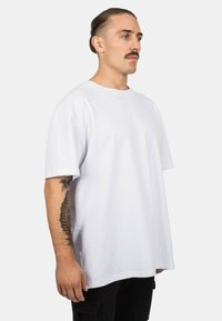 Biela bavlnená tričko s voľným strihom, krátkymi rukávmi a okrúhlym výstrihom, zobrazené na osobe v profile na jednoliatom pozadí.