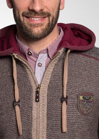 Brauner Strick-Hoodie mit kastanienbraunen Akzenten, beigem Reißverschluss und gesticktem Patch. Darunter ist ein kariertes rot-weißes Hemd sichtbar.