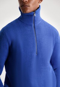 Pulover albastru ajustat dintr-un material tricotat texturat, având un guler înalt și închidere cu fermoar pe jumătate. Designul ribel adaugă interes vizual.