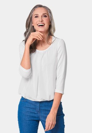 Blusa bianca a maniche lunghe con scollatura rotonda e dettagli plissettati, abbinata a jeans in denim blu. La modella ha capelli grigi lunghi e mossi.