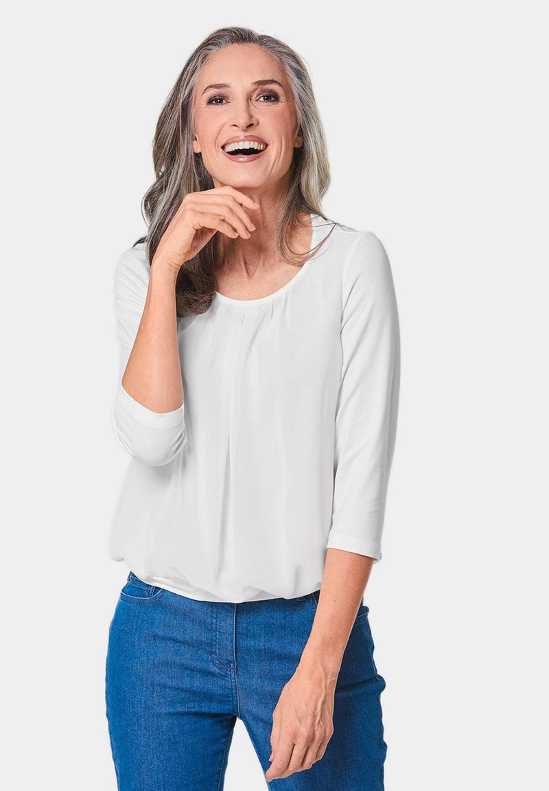 Weiße Bluse mit langen Ärmeln, rundem Ausschnitt und plissierten Details, kombiniert mit blauen Jeans. Das Model hat lange, wellige graue Haare.
