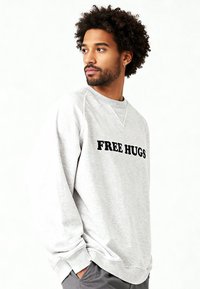 Sweat-shirt gris avec texte noir "FREE HUGS". Comprend des poignets et un col côtelés. Le matériau semble doux avec une coupe décontractée.